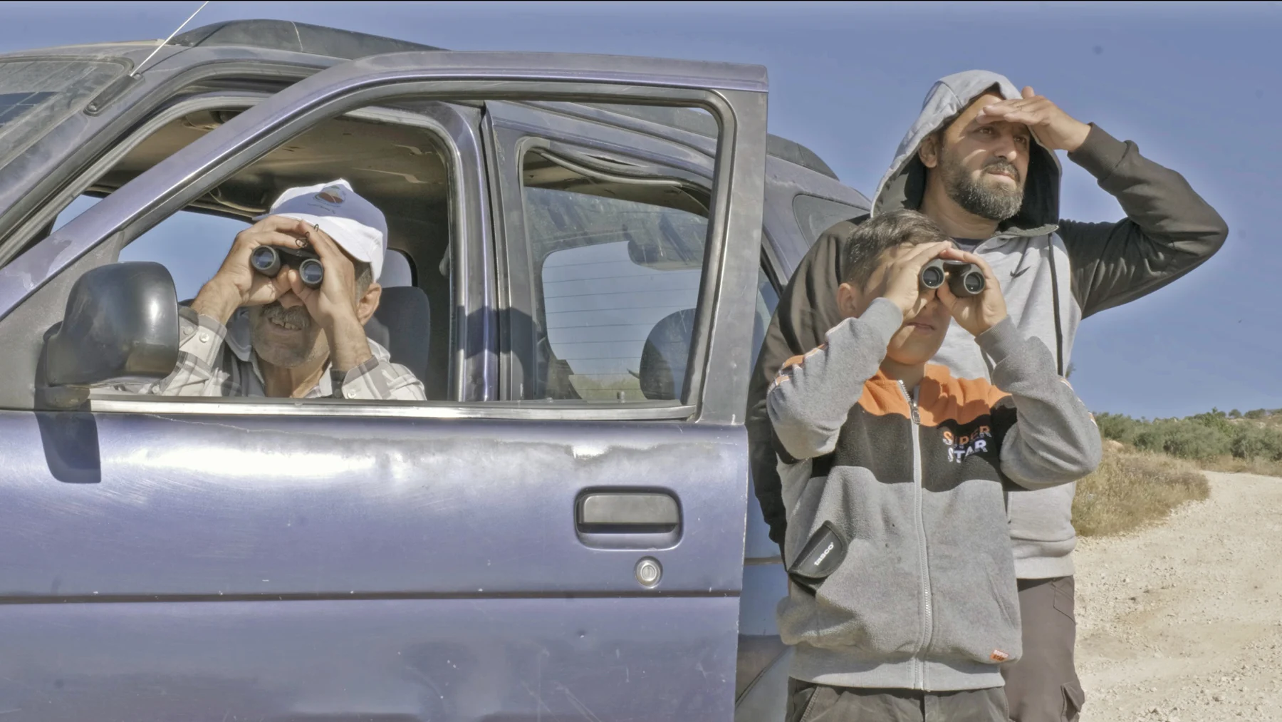 Two people look through binoculars beside a vehicle.